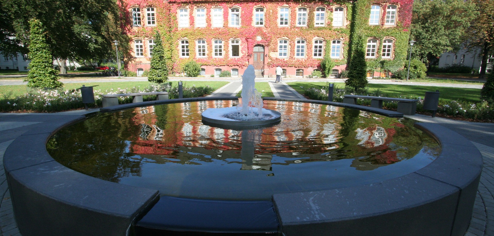 Eine Mann mit Rollator läuft auf das, weinlaubbewachsene, Verwaltungsgebäude zu, Im Vordergrund ein Brunnen. Im Wasser spiegelt sich die Fassade