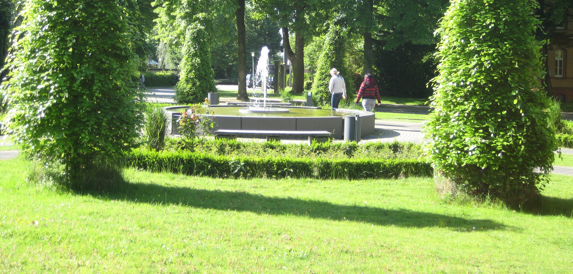 Zwei Frauen gehen an einem sprudelnden Brunnen, der von grünen Bäumen umgeben ist, vorbei. Sie wenden der Kamera den Rücken zu