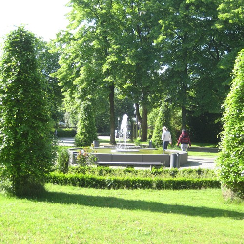 Ein sprudelnder Brunnen umgeben von Grün. Zwei Person laufen an dem Brunnen vorbei.