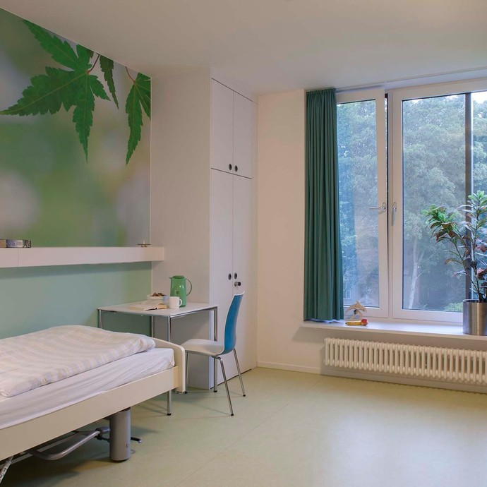 In einem Zimmer steht links, ein Krankenhausbett, dahinter ein Tisch mit weißer Tischplatte, rechts davon ein großes Fenster (vergrößerte Bildansicht wird geöffnet)