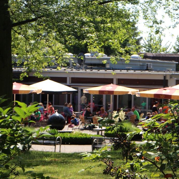 Gäste des Cafés im Sommer auf einer Terrasse mit einem schwarzen, kugelförmigenBrunnen unter Sonnenschirmen. Im Vordergrund grüne Büsche und Pflanzen (vergrößerte Bildansicht wird geöffnet)
