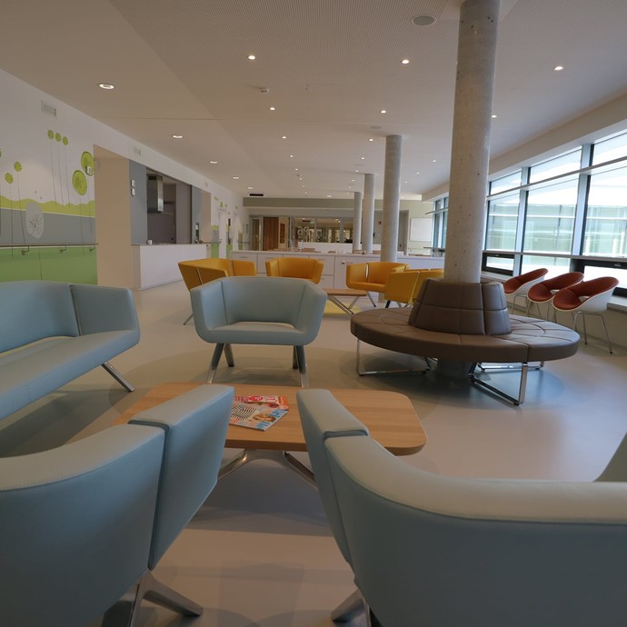 Ein großer, lichtdurchfluteten Raum. It modernen Sitzmöbeln und kleinen Couchtischen, rechts ein großes, bodentiefes Fenster (vergrößerte Bildansicht wird geöffnet)