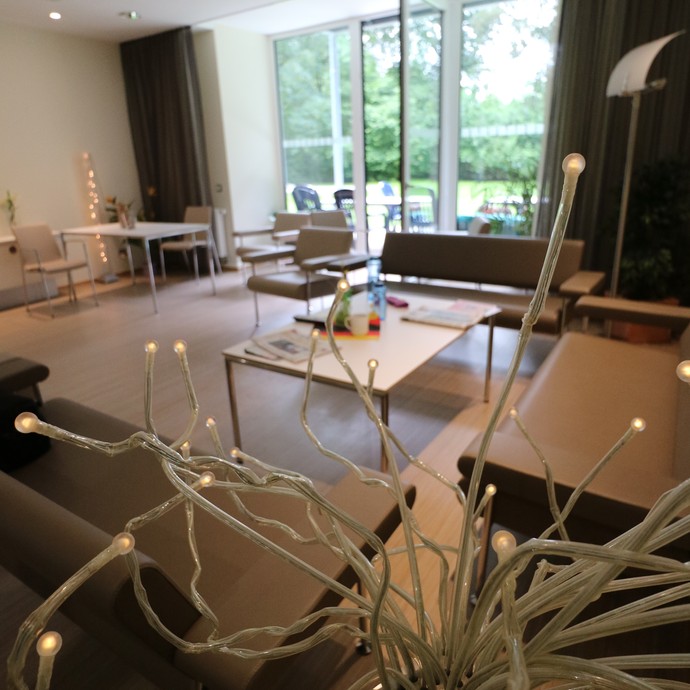 Blick in einen Augfenthaltsraum. Im Vordergrund eine moderne Lampe, mehrere moderne Sitzgelegenheiten, im Hintergrund bodentiefe Fenster, geöffnet (vergrößerte Bildansicht wird geöffnet)
