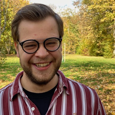 Ein junger Mann mit Brille und Bart steht in einem Park und lächelt freundlich und gewinnend in die Kamera