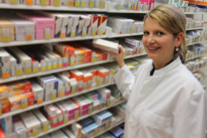 Eine junge Frau in weißem Kittel zieht aus einem Medikamentenregal eine Packung it Tabletten heraus
