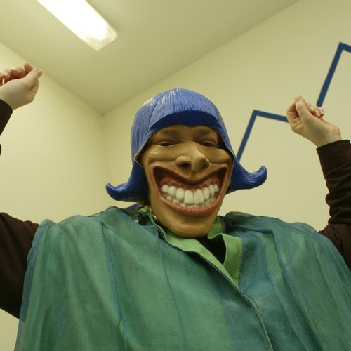 Ein Mann schaut in die Kamera. Er trägt eine lachende Maske und hebt dabei beide Arme