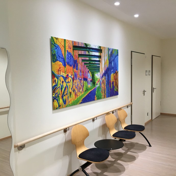Eine hölzerne Sitzbank steht vor einer Wand mit einer grellbunten Fotografie, im Hintergrund zwei geschlssene Türen (vergrößerte Bildansicht wird geöffnet)
