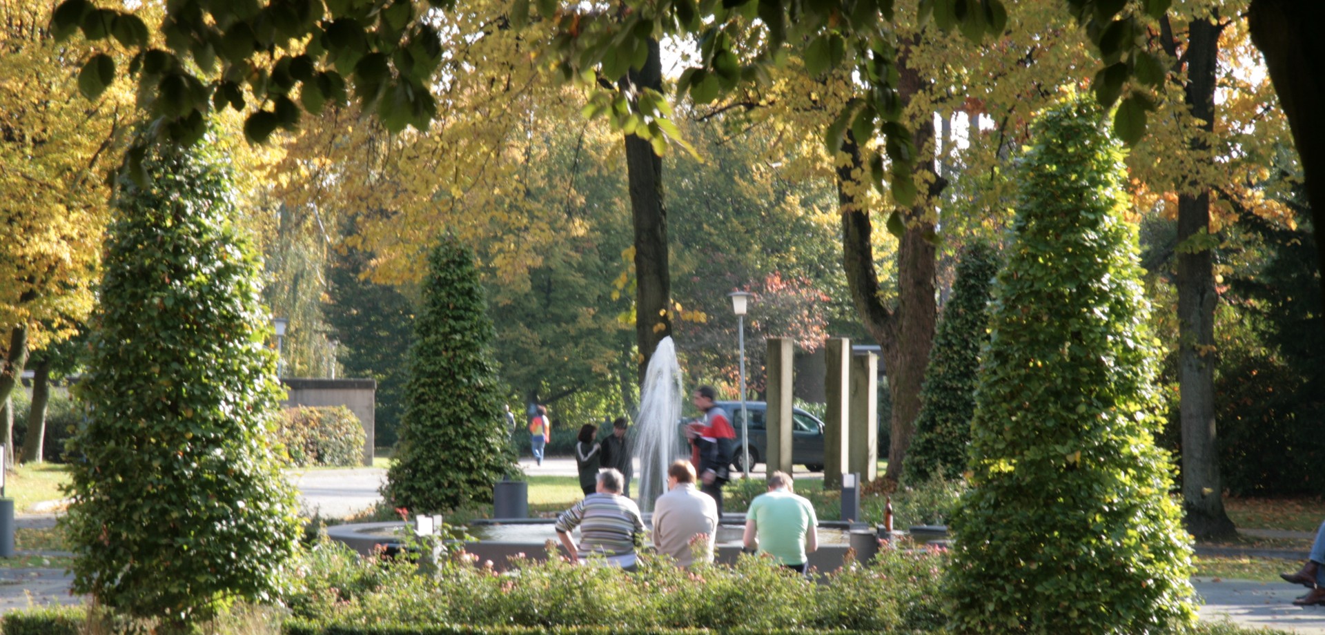 Auf einer Bank sitzen mehrere Personen. Dahinter gehen weitere Personen spazieren