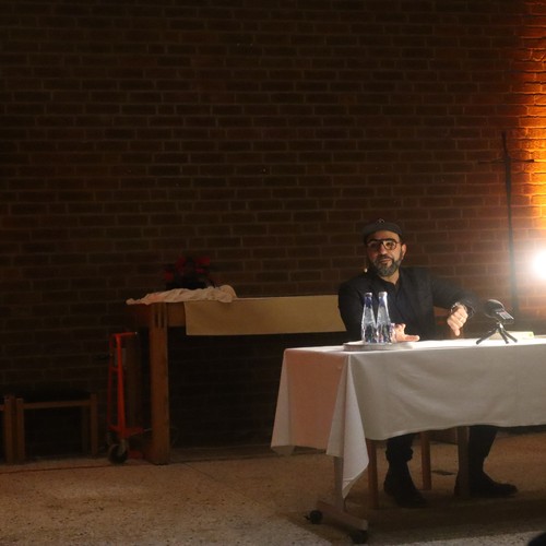 Ein Mann sitzt an einem Tisch in einem abgedunkelten Raum. Im Hintergrund rotes Akzentlicht, welches die Wände beleuchtet