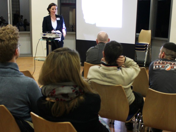 Eine Frau steht vor einer Leinwand. Vor ihr sitzen mehrere Zuhörerinnen und Zuhörer