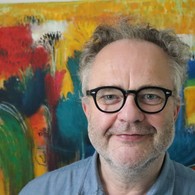 Ein Mann mit dunkel gerahmter Brille steht vor einem Gemälde und schautfreundlich in die Kamera Ein Mann mit dunkel gerahmter Brille steht vor einem Gemälde und schautfreundlich in die Kamera
