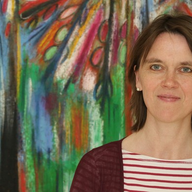 Eine Frau steht vor einem  bunten abstrakten Gemälde und lächelt in die Kamera