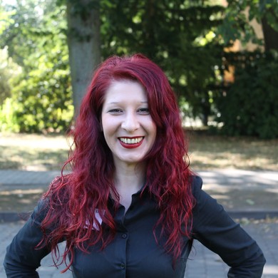 Ein rothaarige Frau steht in einem park und lächelt freundlich in die Kamera