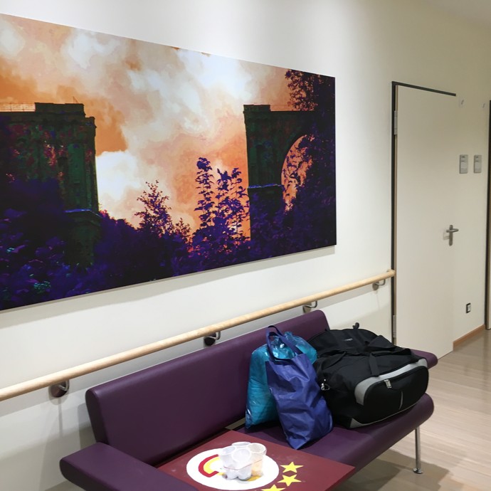 .Eine lilafarbene Sitzbank auf der Gepäck und Einkaufstüten liegen. An der Wand darüber ein lilafarbenes Foto welches eine zerstörte Brücke darstellt. (öffnet vergrößerte Bildansicht)