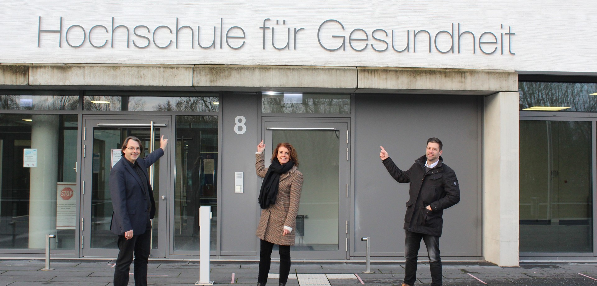 Eine Frau und zwei Männer zeigen auf eine Schriftzug. Hochschule für die Gesundheit