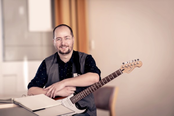 Ein Mann mit Kinnbart lächelt in die Kamera. Er hält eine E-Gitarre im Arm