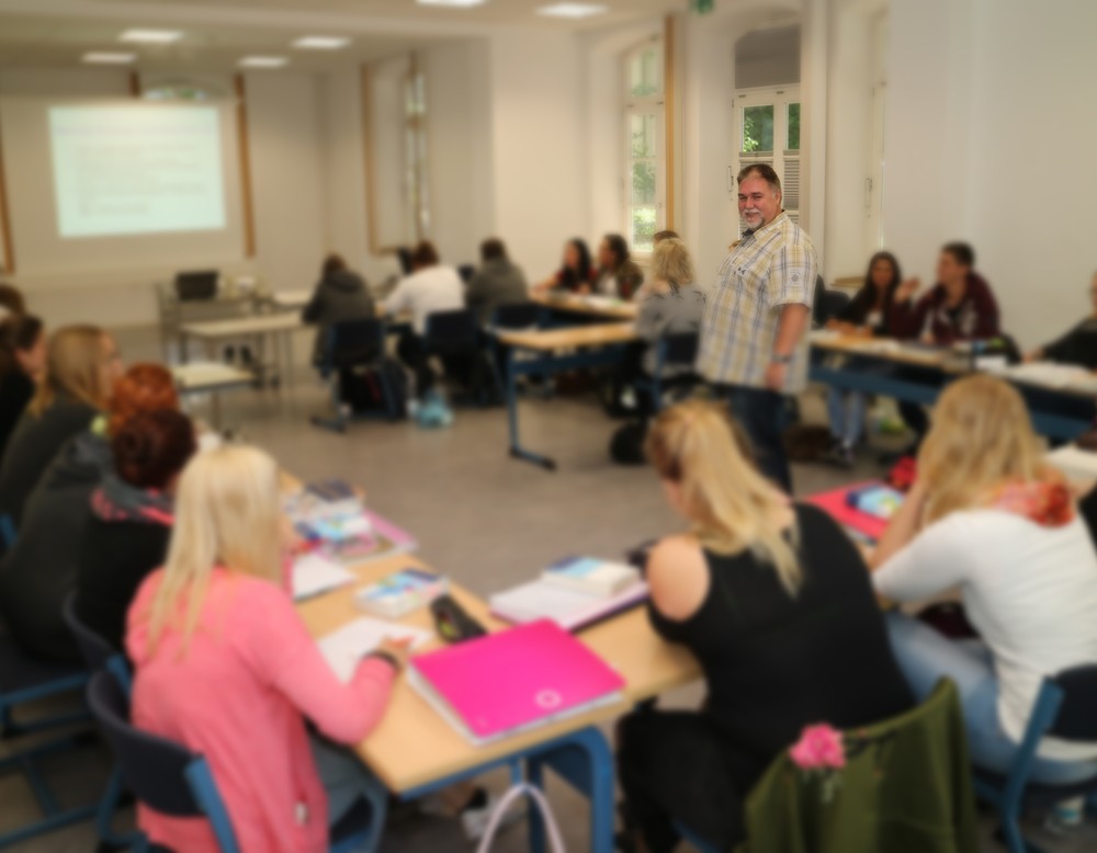 Auszubildende arbeiten an Tischen. In der Mitte der beaufsichtigende Lehrer