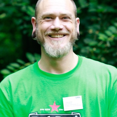 Ein junger, bärtiger Mann  it einem grünen T-Shirt bekleidet lächelt gewinnend  in die Kamera