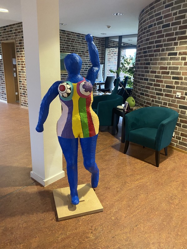 Bunte Frauenskulptur aus Pappmaschee im Innenraum des Tagesklinik Bergkamen. Dahinter zwei einladende, gemütliche Sessel