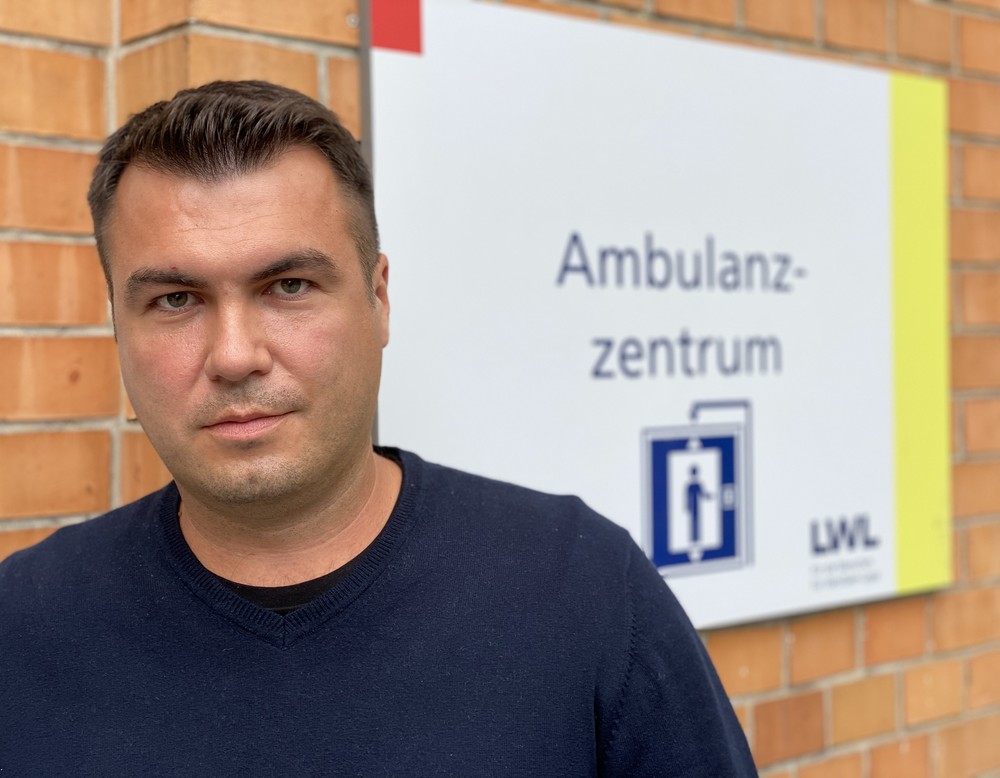 Ein Mann mit kurzem Haarschnitt steht vor einem Praxisschild mit der Aufschr8ft AmbulanzZentrum