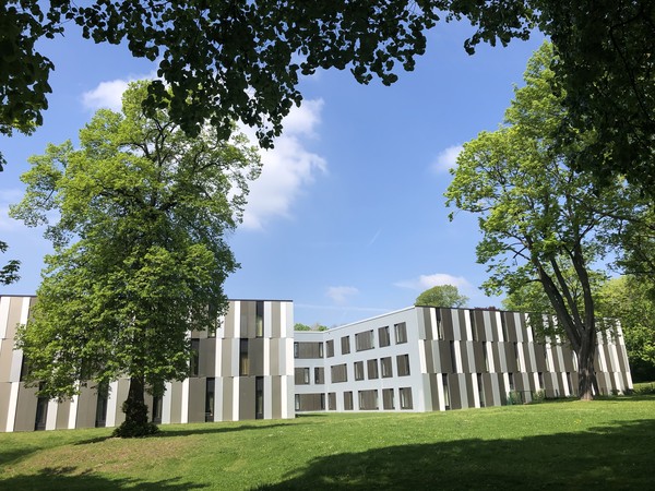 Ein Gebäude mit moderner Fassade. Im Vordergrund eine von Bäumen begrenzte Wiese