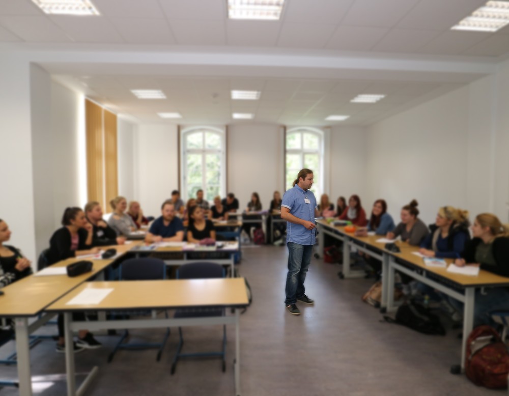 Ein Lehrender steht in einem Klassenraum vor seinen Auszubildenden, die seinen Worten aufmerksam folgen.
