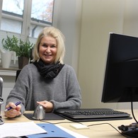Ein blonde Frau sitzt an ihrem Schreibtisch und lächelt gewinnend in die Kamera