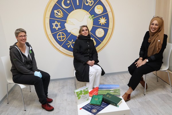 Drei Frauen sitzen in einem hellen Raum. Hinter Ihnen eine Grafik, welche Religionsymbole zeigt,die sich kreisförmnigum eine Friedenstaube gruppieren